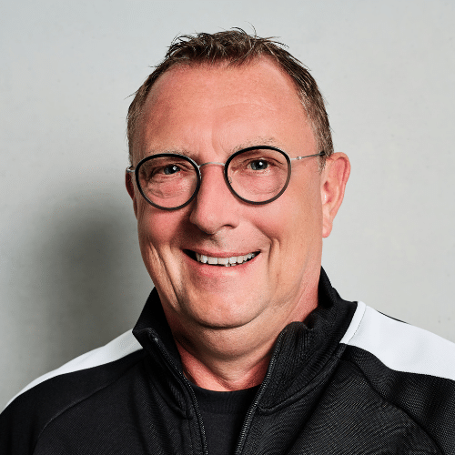 Porträt eines lächelnden Mannes mit Brille, in sportlicher Kleidung, vor einem neutralen Hintergrund.