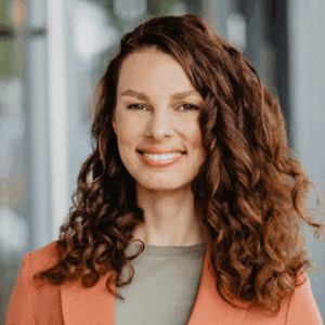 Porträt einer lächelnden Frau mit lockigem, braunem Haar, die einen orangefarbenen Blazer trägt.