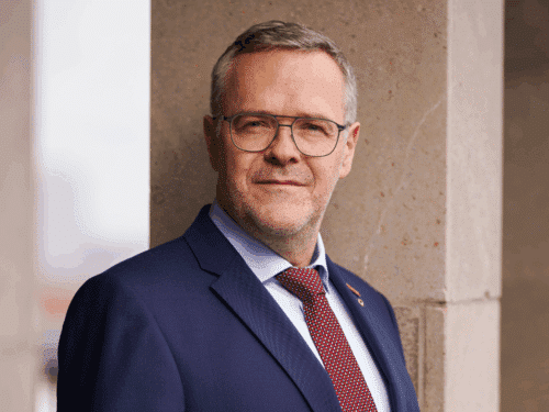 Mann in Anzug mit Brille, lächelt leicht, steht an einer Säule, unscharfer Hintergrund.