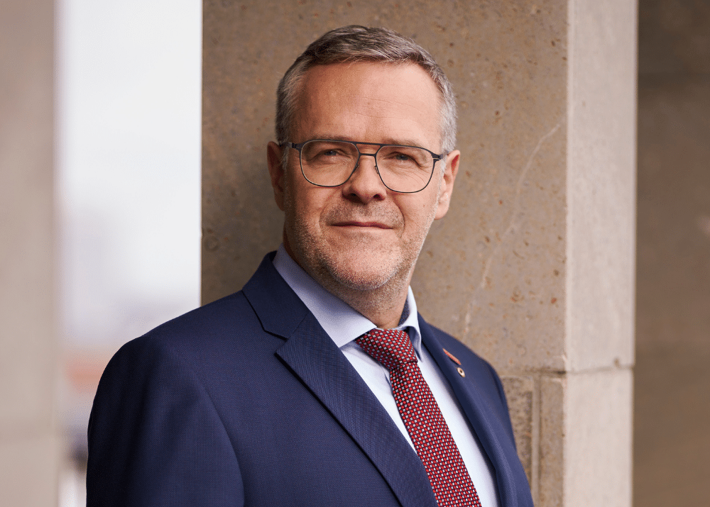 Mann in Anzug mit Brille, lächelt leicht, steht an einer Säule, unscharfer Hintergrund.