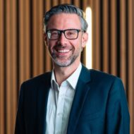 Mann mit Brille und Bart in Anzug, lächelt vor einem Hintergrund aus Holzlamellen und indirekter Beleuchtung.