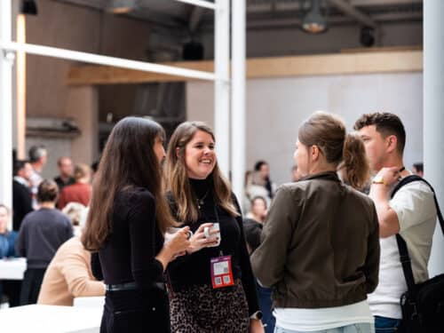 Vier Personen unterhalten sich in einem hellen, modernen Raum mit vielen weiteren Menschen im Hintergrund.