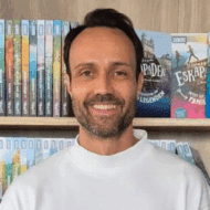 Mann mit braunen Haaren und Bart, trägt einen weißen Pullover, lächelt vor einem Bücherregal.