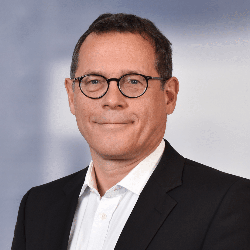 Porträt eines Mannes mit Brille, schwarzem Anzug und weißem Hemd, lächelnd vor unscharfem Hintergrund.