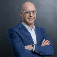 Mann mit Glatze und Brille, lächelt in einem blauen Anzug vor einem dunklen Hintergrund.