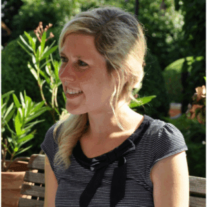 Lächelnde Frau mit blonden Haaren, gestreiftem Shirt und schwarzem Kragen, sitzt im Freien zwischen Pflanzen.