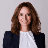 Porträt einer lächelnden Frau mit braunen Haaren, in einem blauen Blazer und weißem Oberteil, vor neutralem Hintergrund.