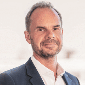 Porträt eines Mannes mit kurzem Haar, Bart und Anzug, lächelnd vor einem hellen Hintergrund.