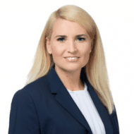 Porträt einer lächelnden Frau mit blonden Haaren, die einen dunkelblauen Blazer und ein weißes Oberteil trägt.