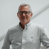 Mann mit Brille und kariertem Hemd steht vor einer hellen Wand.