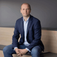 Mann in Anzug sitzt auf einer Bank, lächelt in die Kamera, neutraler Hintergrund.