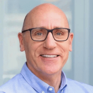 Lächelnder Mann mit Brille und kurz geschorenem Haar, trägt ein hellblaues Hemd, vor einem hellen Hintergrund.