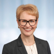 Porträt einer lächelnden Frau mit blonden Haaren, Brille und grauem Blazer, vor unscharfem Hintergrund.