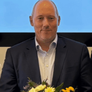 Mann in Anzug mit Blumenstrauß, lächelt vor einem hellen Hintergrund.