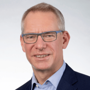 Porträt eines Mannes mit Brille, grauen Haaren und Anzug, vor einem hellen, einfarbigen Hintergrund.