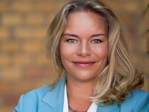 Porträt einer lächelnden Frau mit blonden Haaren, in einem hellblauen Blazer vor einer unscharfen, gemusterten Wand.