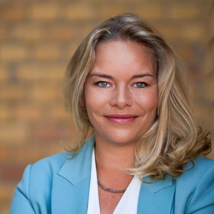 Porträt einer lächelnden Frau mit blonden Haaren, in einem hellblauen Blazer vor einer unscharfen, gemusterten Wand.