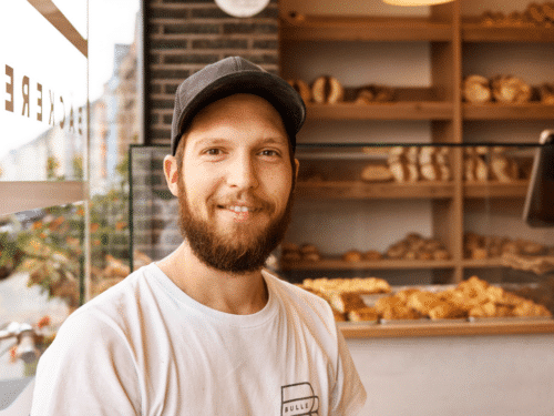 Lächelnder Bäcker mit Bart und Mütze vor einer Vitrine mit frischem Gebäck in einer Bäckerei.