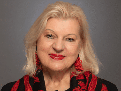 Porträt einer lächelnden Frau mit blonden Haaren, roten Ohrringen und einem schwarzen Schal mit rotem Muster.