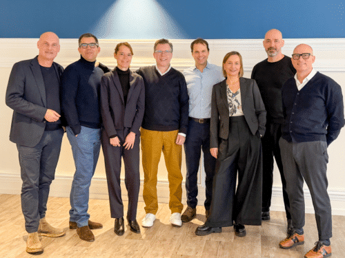 Gruppenfoto von acht Personen in formeller Kleidung, stehend vor einer blauen Wand mit heller Beleuchtung.