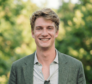 Lächelnder junger Mann mit blonden Haaren, in einem grauen Blazer, vor einer grünen, verschwommenen Hintergrundlandschaft.