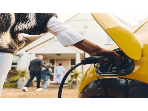 Eine Person steckt ein Ladekabel in ein gelbes Elektroauto vor einem Haus, während andere Personen im Hintergrund stehen.