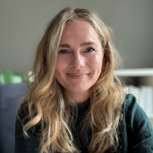 Lächelnde Frau mit langen, blonden Haaren, sitzend vor einem neutralen Hintergrund.