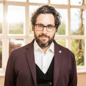 Mann mit lockigem Haar und Brille, trägt einen dunklen Blazer und steht vor einem Fenster mit natürlichem Licht.