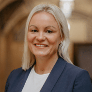 Porträt einer lächelnden Frau mit blonden Haaren, in einem blauen Blazer, vor einem neutralen Hintergrund.