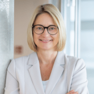 Porträt einer lächelnden Frau mit blonden Haaren und Brille, die in einem hellen Anzug vor einem Fenster steht.