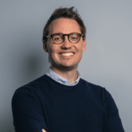 Lächelnder Mann mit Brille, in einem blauen Pullover, vor einem grauen Hintergrund.