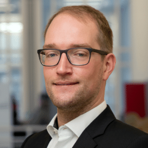 Porträt eines Mannes mit Brille, in Anzug und Hemd, lächelnd vor einem hellen, unscharfen Hintergrund.