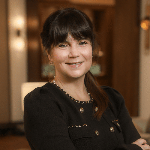 Porträt einer lächelnden Frau mit langen, dunklen Haaren, die in einem eleganten Raum steht.