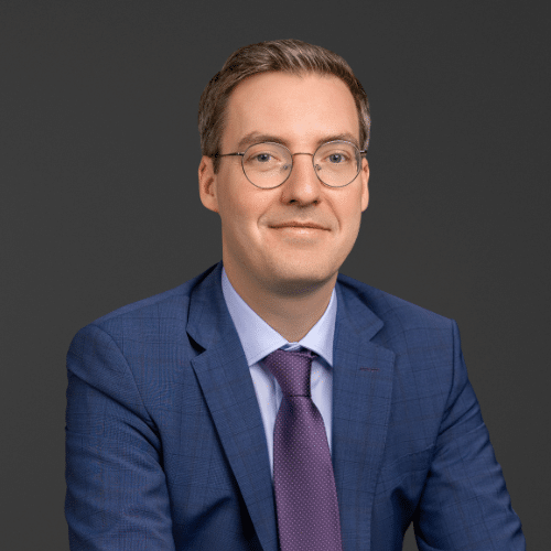 Porträt eines Mannes mit Brille, Anzug und Krawatte, lächelnd vor einem grauen Hintergrund.