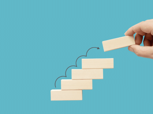 Hand platziert einen Holzblock auf einer Treppe aus Holzblöcken vor einem blauen Hintergrund.