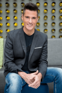 Mann mit dunklem Anzug und T-Shirt sitzt lächelnd auf einem Hocker vor einer modernen Wand mit runden, gelben Elementen.