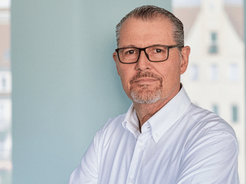 Mann mit Brille und Hemd, steht mit verschränkten Armen vor einem hellen Hintergrund.