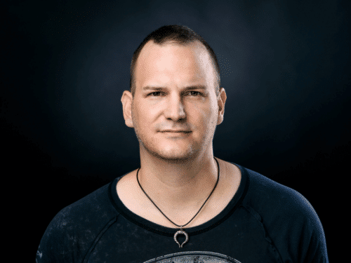 Porträt eines Mannes mit kurzem Haar, neutralem Gesichtsausdruck, schwarzem T-Shirt und Halskette vor dunklem Hintergrund.