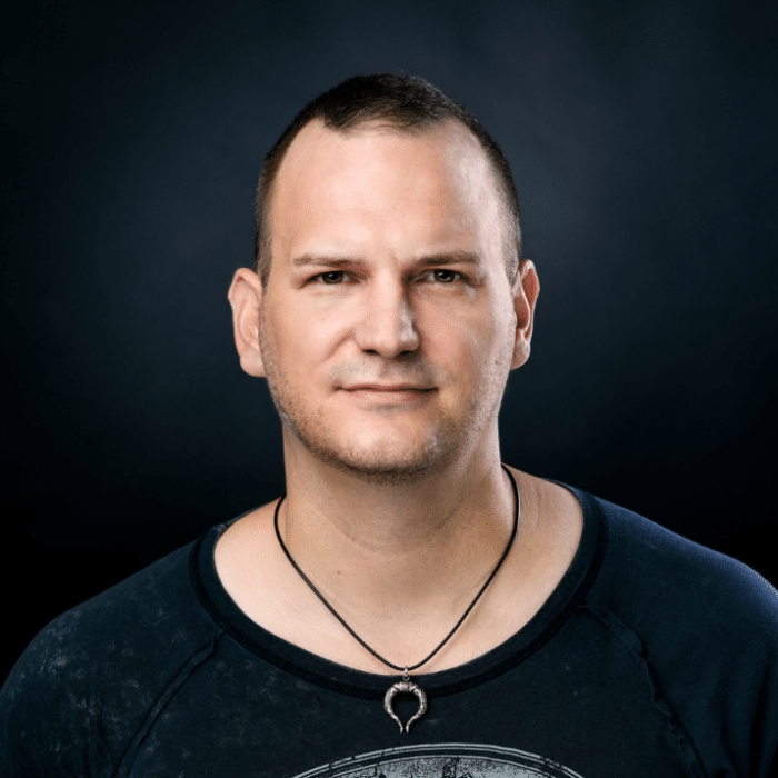 Porträt eines Mannes mit kurzem Haar, neutralem Gesichtsausdruck, schwarzem T-Shirt und Halskette vor dunklem Hintergrund.