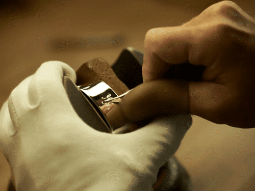 Eine Hand in einem weißen Handschuh graviert ein glänzendes Metallstück auf einem Holzuntergrund.