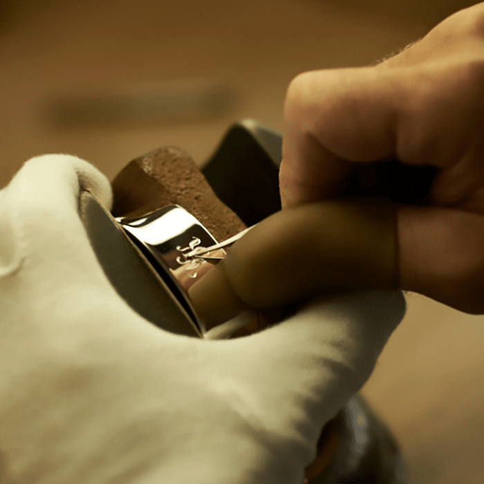 Eine Hand in einem weißen Handschuh graviert ein glänzendes Metallstück auf einem Holzuntergrund.