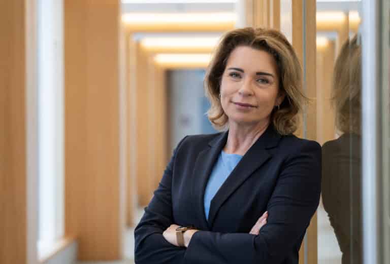 Frau mit lockigem Haar in einem Blazer steht in einem modernen Flur mit Holz- und Glaselementen.