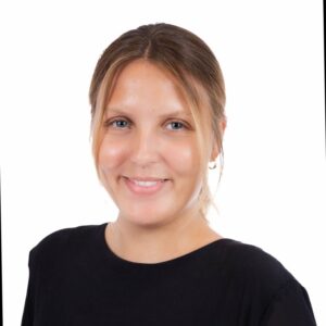 Porträt einer lächelnden Frau mit blonden Haaren, die ein schwarzes Oberteil trägt, vor einem weißen Hintergrund.