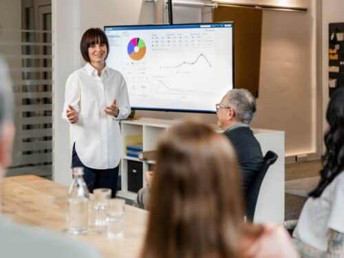 Frau in weißem Hemd präsentiert Daten auf einem Bildschirm vor einer Gruppe von Zuhörern in einem modernen Büro.