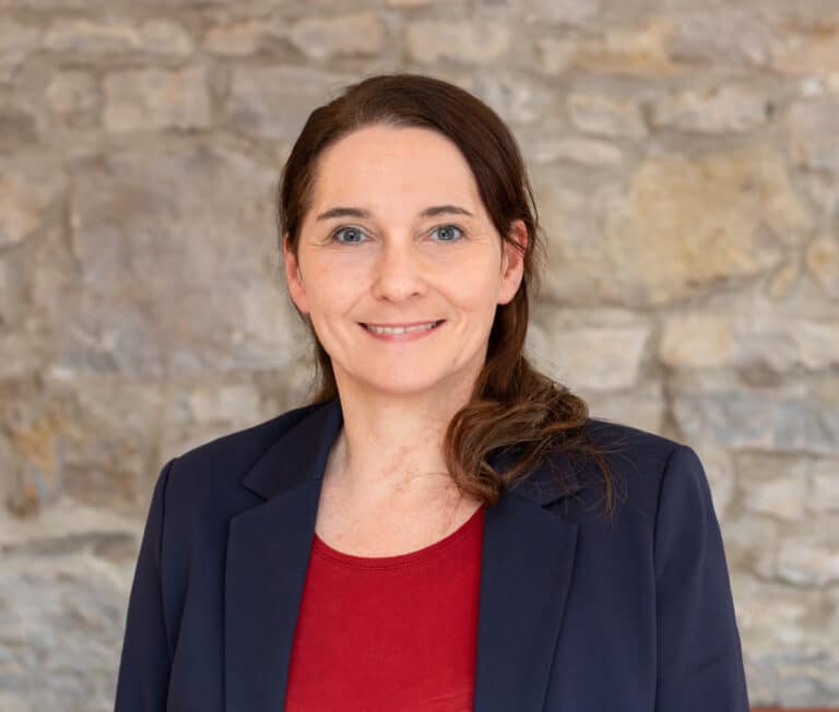 Porträt einer Frau mit langen, braunen Haaren, die in einem blauen Blazer und rotem Oberteil vor einer Steinmauer steht.