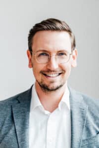 Mann mit Brille und lächelndem Gesicht, trägt einen grauen Blazer und ein weißes Hemd, vor neutralem Hintergrund.