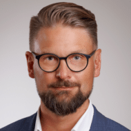 Porträt eines Mannes mit Brille, kurzem Haar und Bart, in einem Anzug vor neutralem Hintergrund.