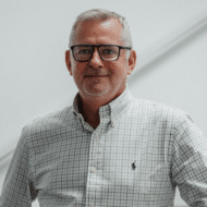 Mann mit Brille und kariertem Hemd steht vor einer hellen Wand.