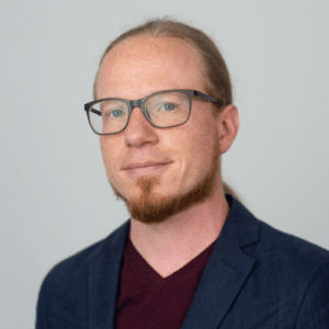 Porträt eines Mannes mit Brille, langen Haaren und Bart, der einen dunkelblauen Blazer und ein rotes Shirt trägt.