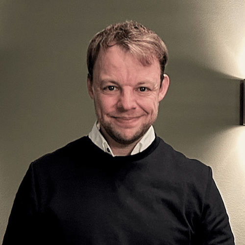 Lächelnde Person in schwarzem Pullover vor einer hellen Wand mit indirekter Beleuchtung.